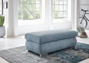 XXL-Hocker in hellblauem Stoff, seitliche Perspektive, in einem hellen Raum mit großen Fenstern und moderner Einrichtung.