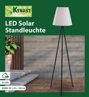 LED Solar Standleuchte mit dreibeinigem Stativ und weißem Lampenschirm, seitliche Perspektive, aufgestellt auf einer Terrasse mit Holzboden und Pflanze im Hintergrund.