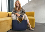 Frau sitzt auf einem gelben Sofa und hält eine große, dunkelblaue Tasche mit runden Griffen. Perspektive von vorne.