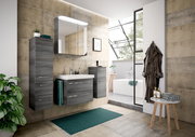 Moderne Badezimmermöbel in grauer Holzoptik, bestehend aus einem Waschtisch mit Spiegel und Schränken, in einer stilvollen Badezimmerumgebung mit Blick auf eine Badewanne und große Fenster.