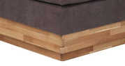 Nahaufnahme der Ecke eines Boxspringbetts mit grauem Stoffbezug und Holzunterbau, Perspektive von unten.