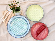 Draufsicht auf ein 12-teiliges Tellerset mit bunten Mustern in Blau, Grün und Rot, arrangiert auf einem Leinentuch mit Besteck und Blumen.