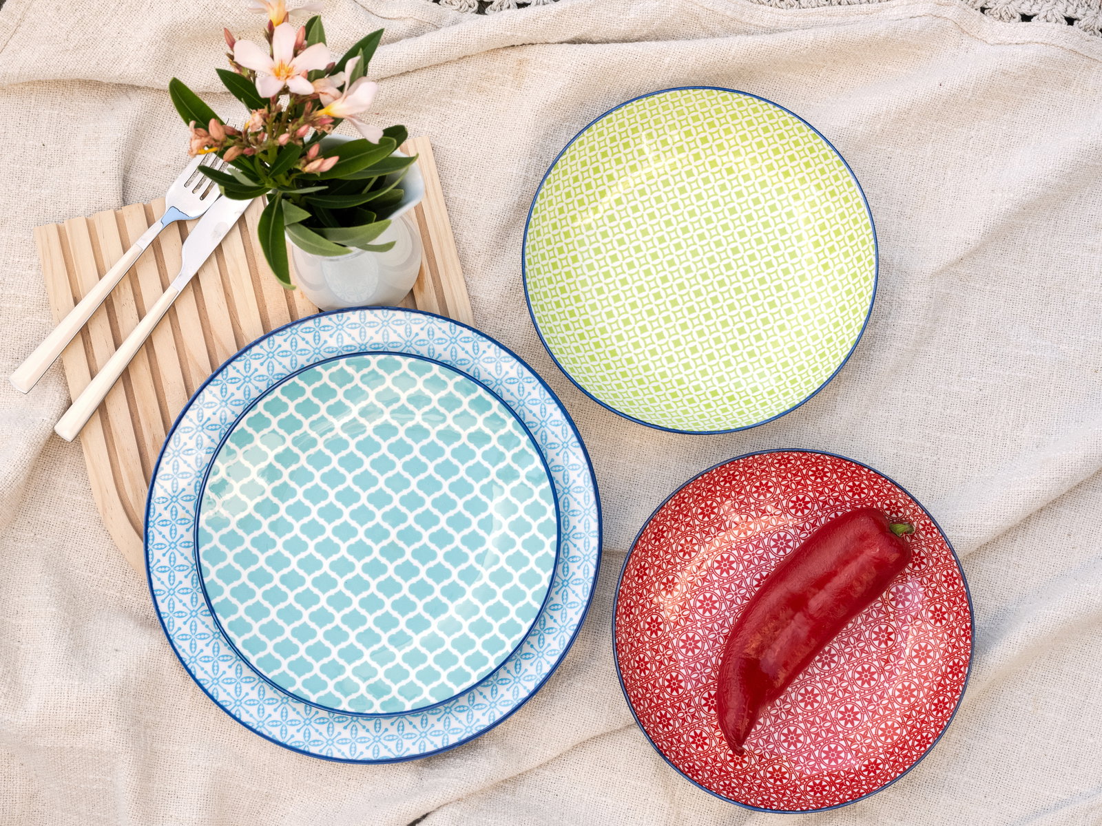 Draufsicht auf ein 12-teiliges Tellerset mit bunten Mustern in Blau, Grün und Rot, arrangiert auf einem Leinentuch mit Besteck und Blumen.