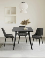 Moderner Esstisch mit schwarzer Tischplatte und vier grauen Stühlen, frontal betrachtet. Über dem Tisch hängt eine stilvolle weiße Lampe, und auf dem Tisch steht eine Vase mit getrockneten Gräsern.