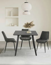 Moderner Esstisch mit schwarzer Tischplatte und vier grauen Stühlen, frontal betrachtet. Über dem Tisch hängt eine stilvolle weiße Lampe, und auf dem Tisch steht eine Vase mit getrockneten Gräsern.