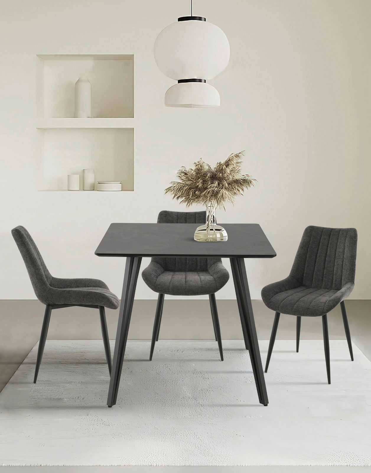 Moderner Esstisch mit schwarzer Tischplatte und vier grauen Stühlen, frontal betrachtet. Über dem Tisch hängt eine stilvolle weiße Lampe, und auf dem Tisch steht eine Vase mit getrockneten Gräsern.