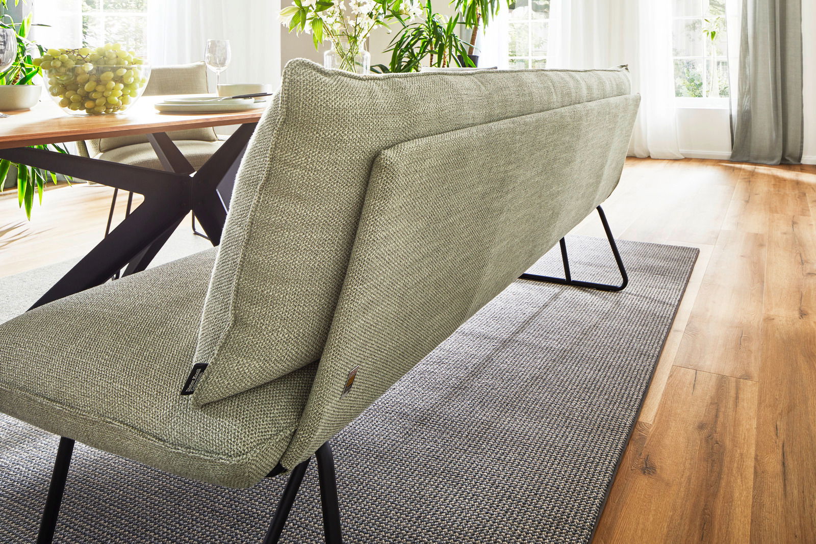 Rückansicht einer modernen, gepolsterten Bank mit Metallbeinen in einem hellen Raum mit Holzboden und einem Esstisch im Hintergrund.