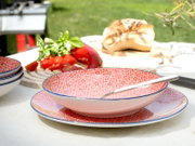 Rosa Keramikschale mit rotem geometrischem Muster und blauem Rand auf passendem Teller, mit Löffel, auf gedecktem Outdoor-Tisch; Perspektive: Nahaufnahme aus leicht schräger Seitenansicht.