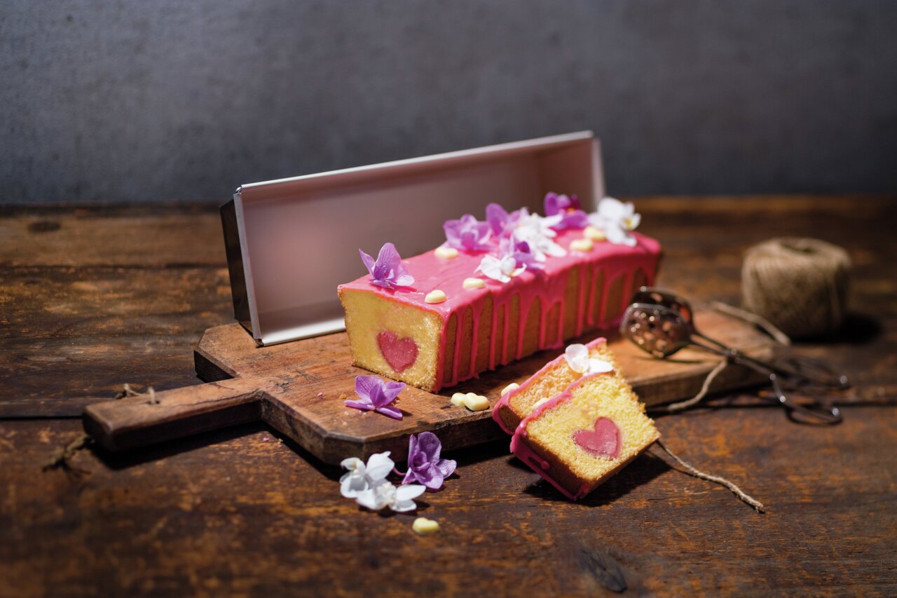 Kastenform 30 cm BackTrend mit einem gebackenen Kuchen, dekoriert mit rosa Glasur und Blumen, auf einem Holzbrett präsentiert. Perspektive von vorne.