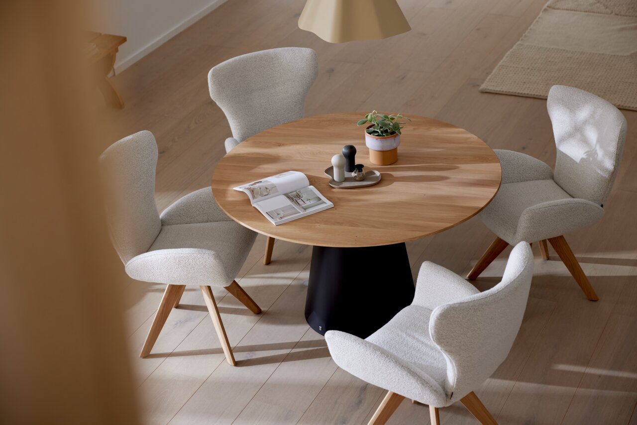 Musterring Esstisch MR JUSTB! Runder Esstisch aus Holz mit schwarzem Sockel, umgeben von vier gepolsterten Armlehnstühlen in einem modernen Esszimmer. Perspektive von oben.