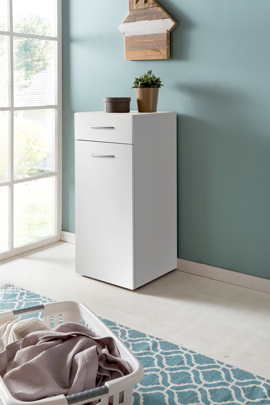 Weiße Wäschekommode mit Schublade und Tür, seitliche Perspektive, dekoriert mit Pflanze und Box, vor mintgrüner Wand.