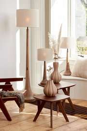 Ein stilvoller Raum mit mehreren Lampen mit beigen Lampenschirmen, die auf einem Holztisch und auf dem Boden stehen. Die Perspektive zeigt die Lampen von der Seite, mit natürlichem Licht, das durch ein Fenster einfällt.