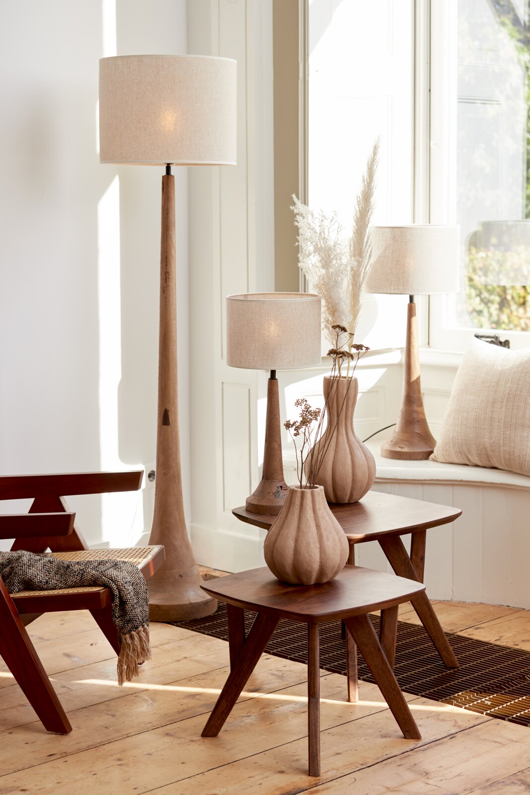Een stijlvolle ruimte met verschillende lampen met beige lampenkappen, die op een houten tafel en op de vloer staan. Het perspectief toont de lampen van opzij, met natuurlijk licht dat door een raam naar binnen valt.