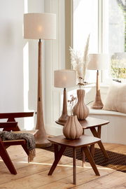 Een stijlvolle ruimte met verschillende lampen met beige lampenkappen, die op een houten tafel en op de vloer staan. Het perspectief toont de lampen van opzij, met natuurlijk licht dat door een raam naar binnen valt.