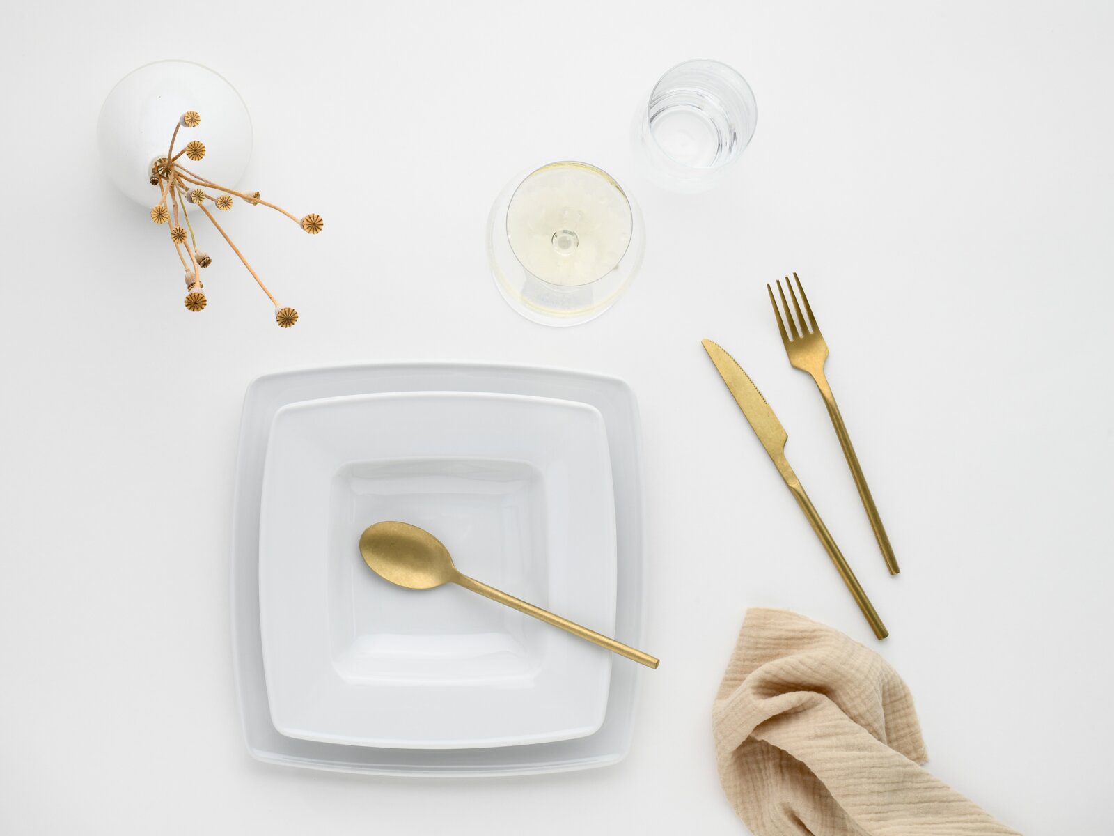 Draufsicht auf minimalistisches Tischgedeck: weißer quadratischer Teller mit goldfarbenem Löffel, daneben goldfarbenes Messer und Gabel, zwei Gläser, beige Serviette und dekorative Trockenblumen auf weißem Hintergrund