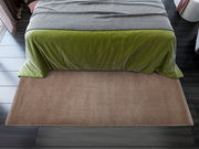 Weiche, beige Matte vor einem Bett mit grünem Überwurf, aus der Vogelperspektive fotografiert.