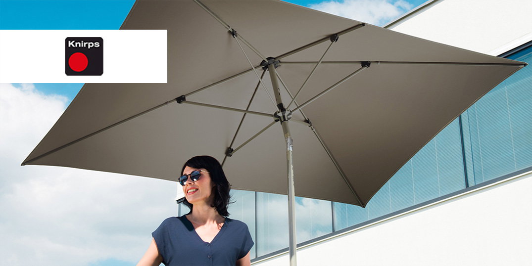 Een vrouw staat onder een grote beige parasol. Het Knirps-logo in de hoek.