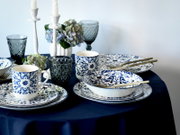 Gedeckter Tisch mit blau-weiß gemustertem Porzellan (Teller, Schalen, Tassen), goldfarbenem Besteck, dunkelblauen Gläsern, weißen Kerzenständern und Hortensien auf dunkelblauer Tischdecke, in seitlicher Perspektive leicht von oben