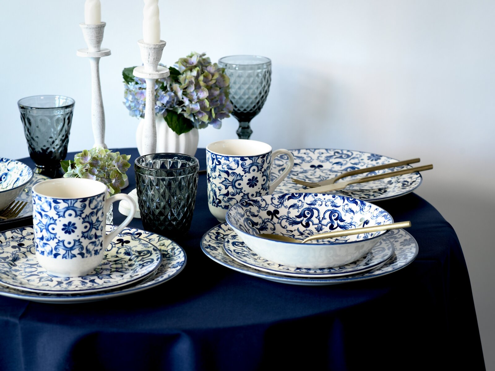 Gedeckter Tisch mit blau-weiß gemustertem Porzellan (Teller, Schalen, Tassen), goldfarbenem Besteck, dunkelblauen Gläsern, weißen Kerzenständern und Hortensien auf dunkelblauer Tischdecke, in seitlicher Perspektive leicht von oben