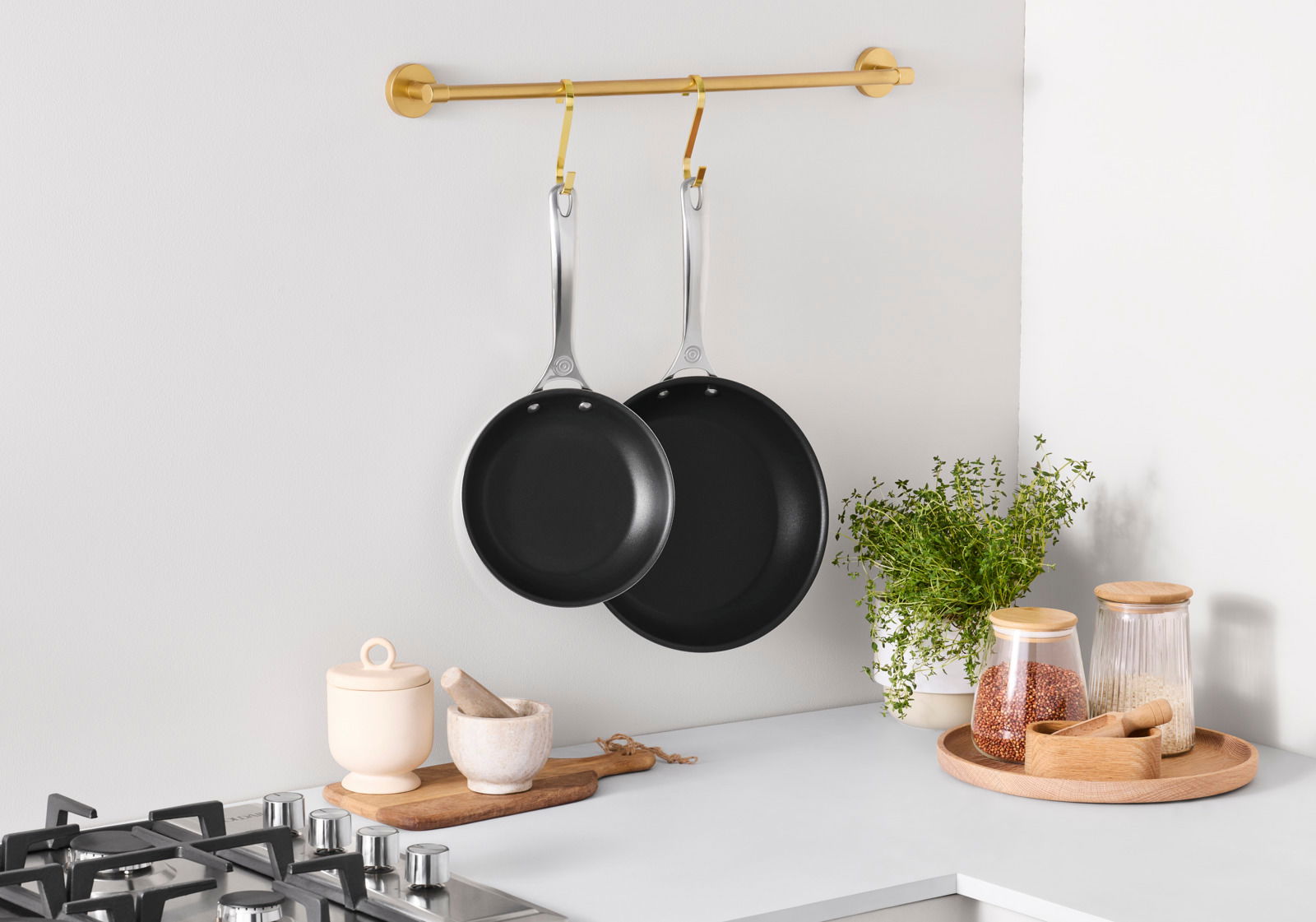 Zwei 3-PLY PLUS Pfannen in den Größen 20cm und 26cm hängen an einer goldenen Stange in einer modernen Küche. Die Pfannen sind aus Edelstahl mit schwarzer Antihaftbeschichtung. Perspektive von vorne.
