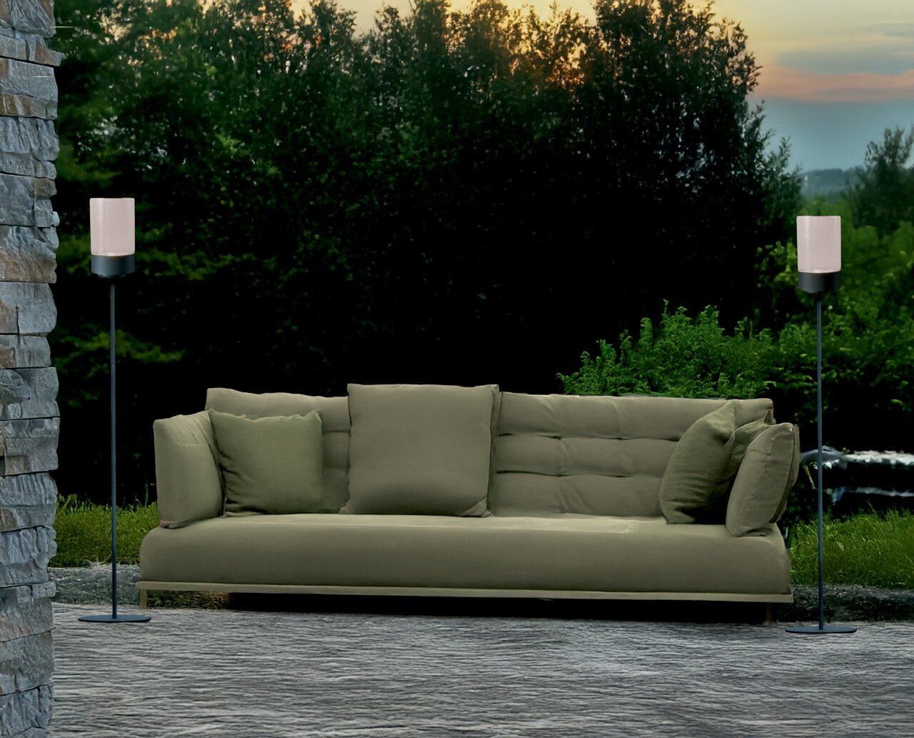 Zwei Akku-Windlichter auf Ständern flankieren ein grünes Sofa im Freien, umgeben von Natur, aus frontaler Perspektive.