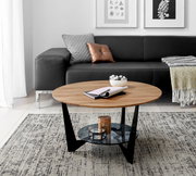 Runder Couchtisch aus Holz mit schwarzem Metallgestell und Glasablage, im Wohnzimmer mit grauem Sofa und dekorativen Kissen, aus seitlicher Perspektive.