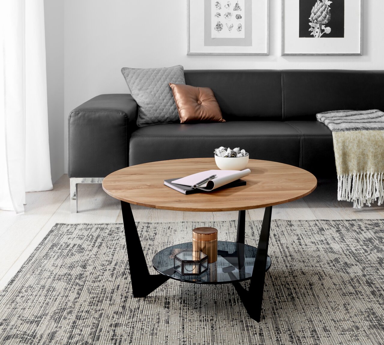 Runder Couchtisch aus Holz mit schwarzem Metallgestell und Glasablage, im Wohnzimmer mit grauem Sofa und dekorativen Kissen, aus seitlicher Perspektive.