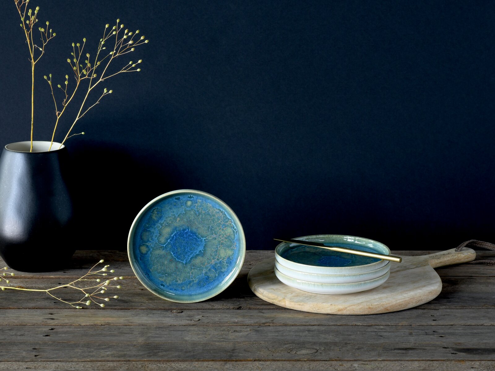 creaTable Brotteller 4-tlg YUKI Blau glasierte Keramikschale mit reaktiver Glasur, frontale Ansicht auf rustikalem Holztisch; daneben gestapelte Schalen auf Holzbrett mit goldenem Löffel und schwarze Vase vor dunkelblauem Hintergrund.