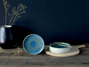 Blau glasierte Keramikschale mit reaktiver Glasur, frontale Ansicht auf rustikalem Holztisch; daneben gestapelte Schalen auf Holzbrett mit goldenem Löffel und schwarze Vase vor dunkelblauem Hintergrund.