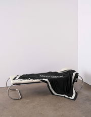 Schwarzes Plaid mit weißem Rand und Schriftzug, drapiert auf einer modernen Liege aus Metall und Leder, seitliche Perspektive.