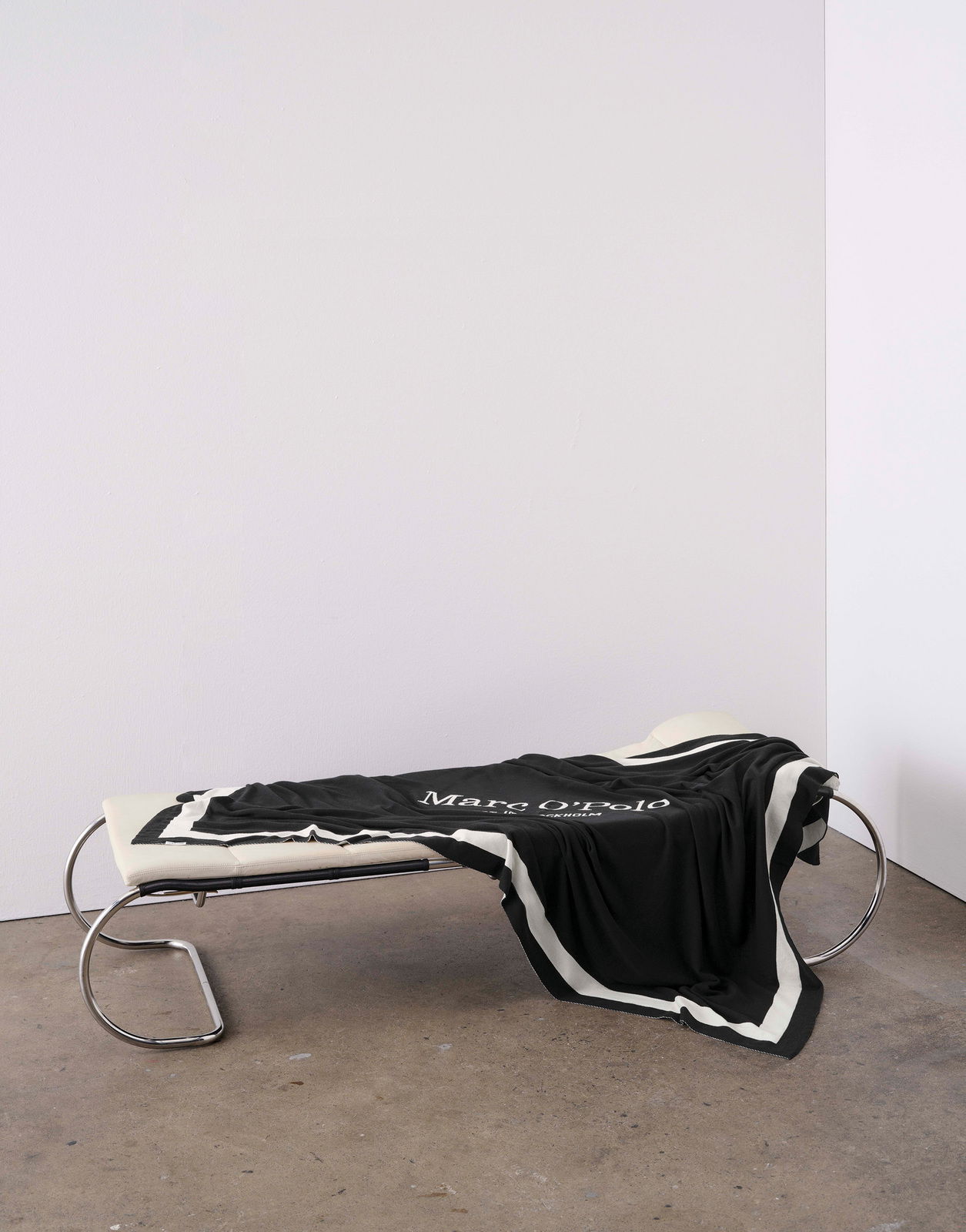 Schwarzes Plaid mit weißem Rand und Schriftzug, drapiert auf einer modernen Liege aus Metall und Leder, seitliche Perspektive.