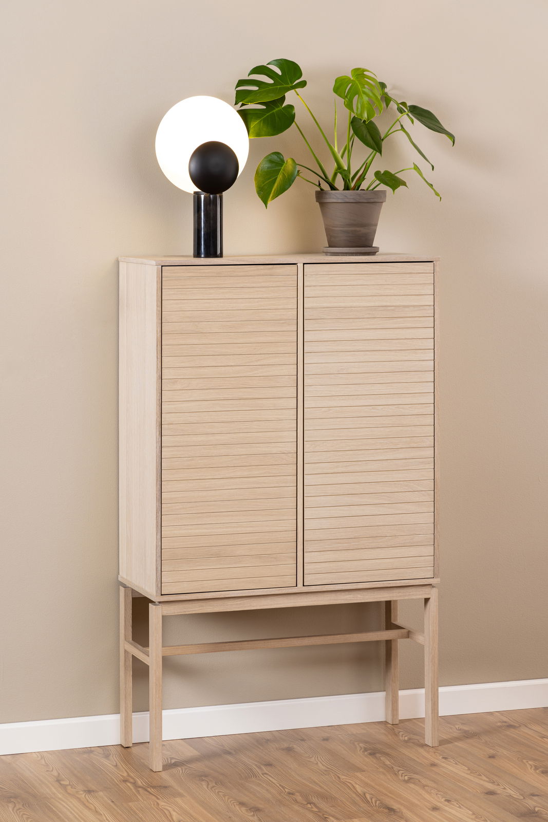 Hochschrank aus hellem Holz mit zwei Türen, darauf eine moderne Lampe und eine grüne Pflanze, seitliche Perspektive