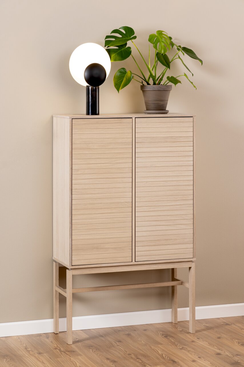 Hochschrank aus hellem Holz mit zwei Türen, darauf eine moderne Lampe und eine grüne Pflanze, seitliche Perspektive