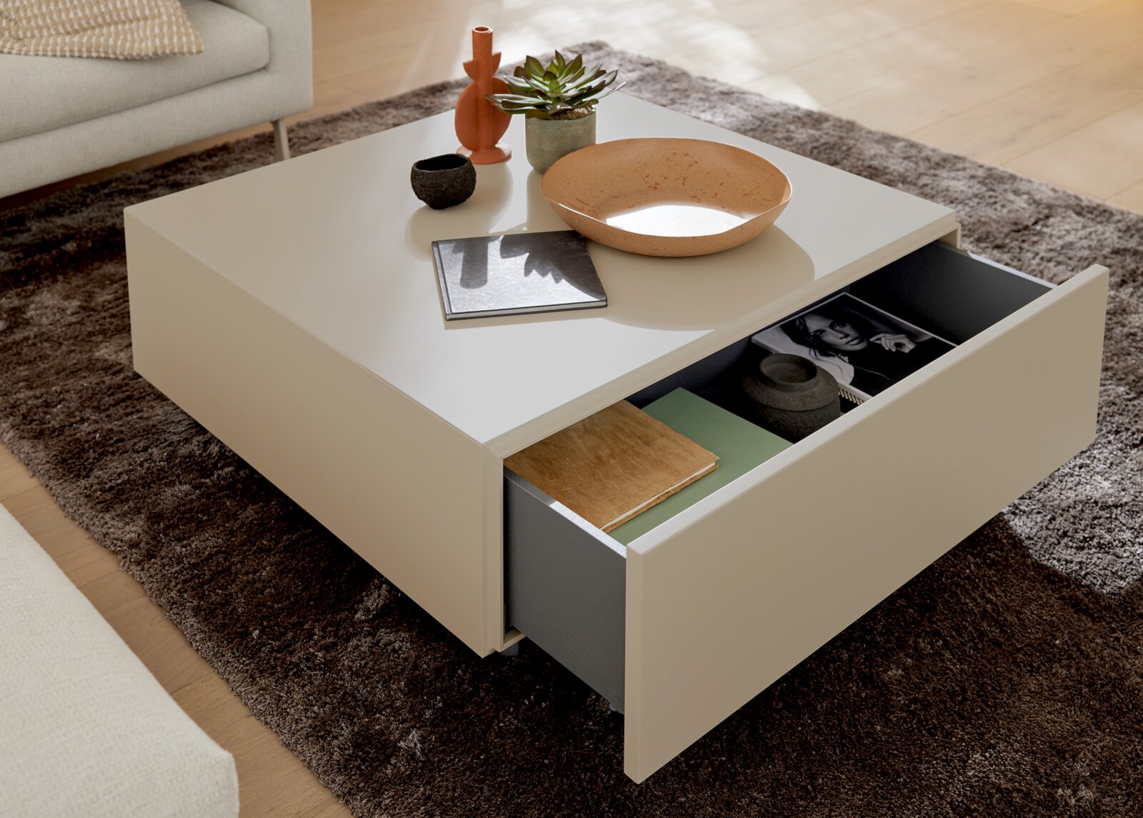 Moderner quadratischer Couchtisch in Cremeweiß mit geöffneter, geräumiger Schublade, dekoriert mit Schale, Pflanze und Wohnaccessoires, aus schräger Perspektive von oben.