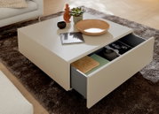 Moderner quadratischer Couchtisch in Cremeweiß mit geöffneter, geräumiger Schublade, dekoriert mit Schale, Pflanze und Wohnaccessoires, aus schräger Perspektive von oben.