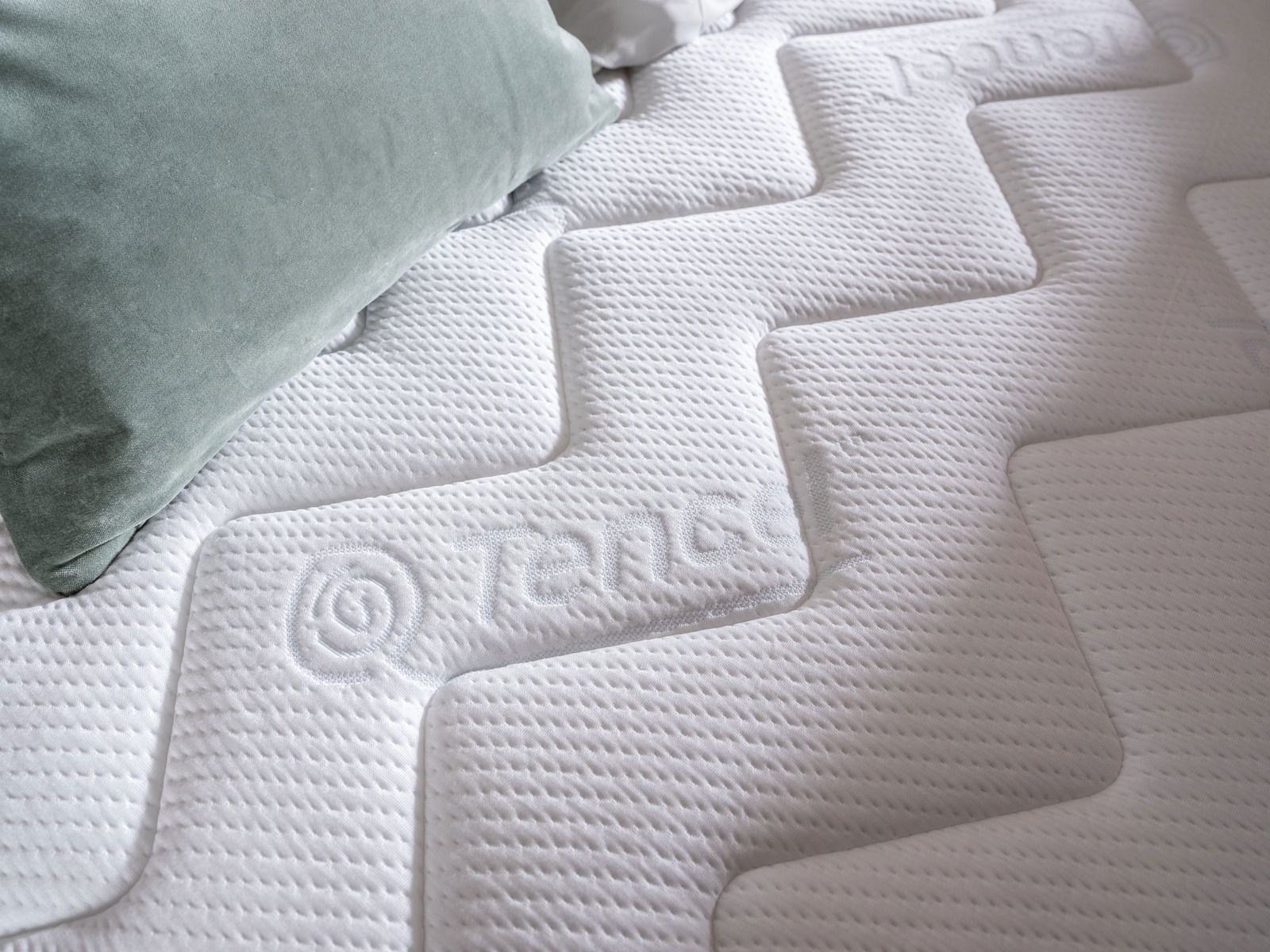 Nahaufnahme einer weißen Matratze mit geprägtem Muster und einem grünen Kissen, von oben fotografiert.