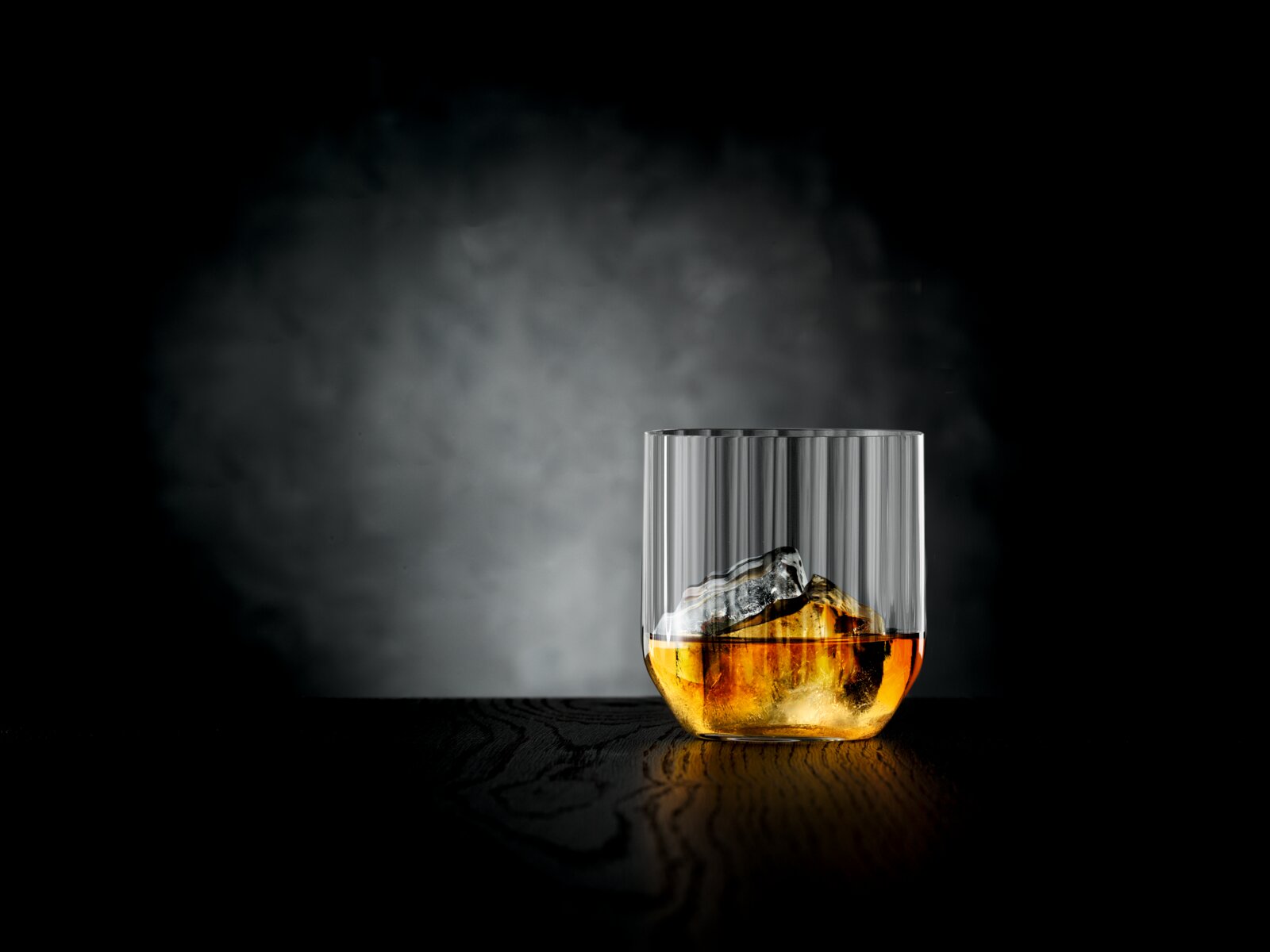 Ein einzelnes Glas mit geriffeltem Design, gefüllt mit einem Getränk und Eiswürfeln, auf einer dunklen Oberfläche vor einem dunklen Hintergrund, seitliche Perspektive.