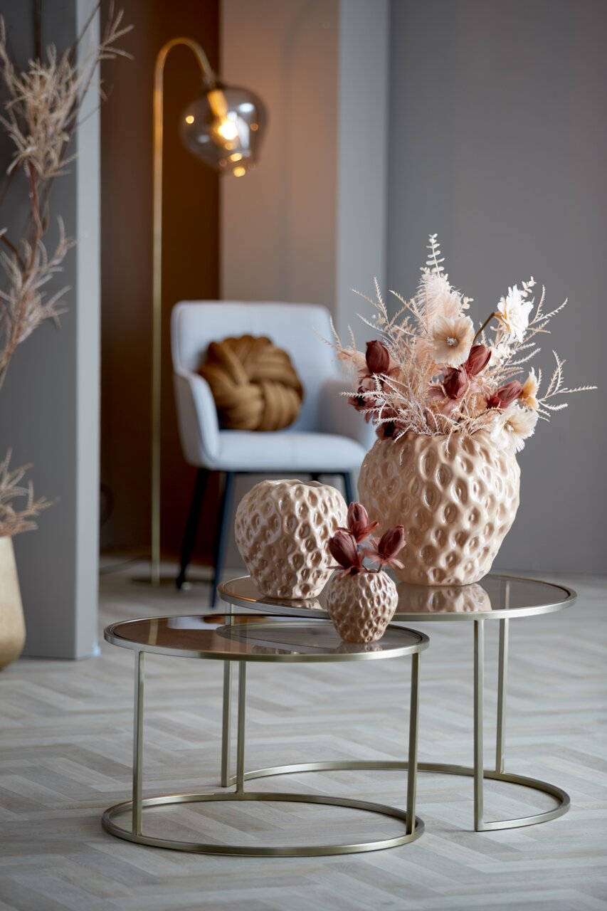 Zwei runde Couchtische mit Glasplatte und goldfarbenem Gestell, dekoriert mit strukturierten Vasen und Trockenblumen, in einem modernen Wohnzimmer mit seitlicher Perspektive.