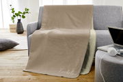Beige Jacquard-Decke 150x200cm auf grauem Sofa, mit Schriftzug 'Kuscheldecke', in einem gemütlichen Wohnzimmer mit Holzboden und Pflanzen im Hintergrund, seitliche Perspektive.