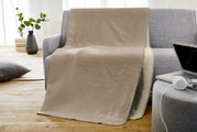 Beige Jacquard-Decke 150x200cm auf grauem Sofa, mit Schriftzug 'Kuscheldecke', in einem gemütlichen Wohnzimmer mit Holzboden und Pflanzen im Hintergrund, seitliche Perspektive.
