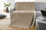 Beige Jacquard-Decke 150x200cm auf grauem Sofa, mit Schriftzug 'Kuscheldecke', in einem gemütlichen Wohnzimmer mit Holzboden und Pflanzen im Hintergrund, seitliche Perspektive.