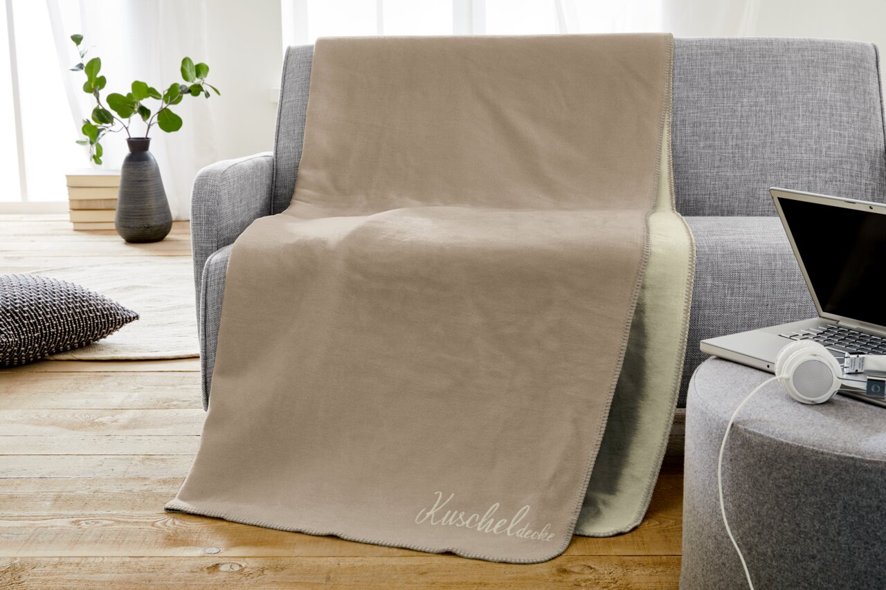 IBENA Jacquard Wohndecke KUSCHELDECKE Beige Jacquard-Decke 150x200cm auf grauem Sofa, mit Schriftzug 'Kuscheldecke', in einem gemütlichen Wohnzimmer mit Holzboden und Pflanzen im Hintergrund, seitliche Perspektive.
