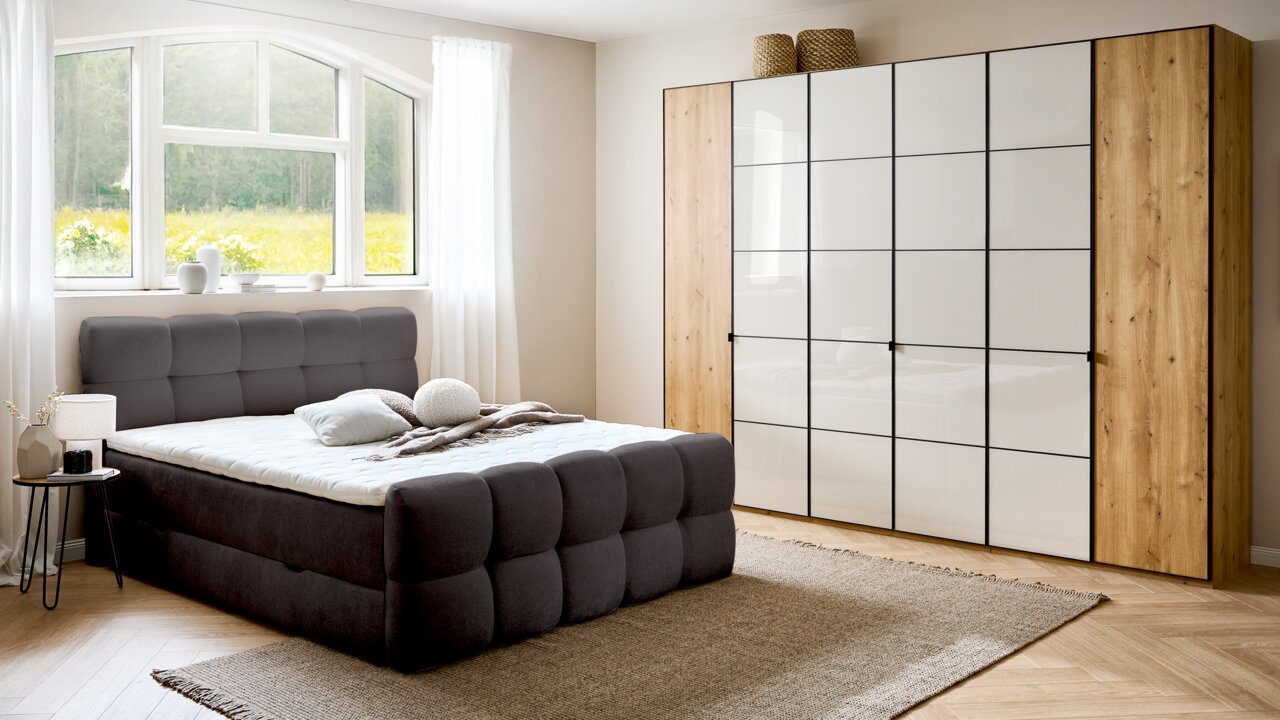 Modernes Boxbett in einem hellen Schlafzimmer mit großem Fenster, grauem gepolstertem Kopfteil und passendem Schrank, seitliche Perspektive.