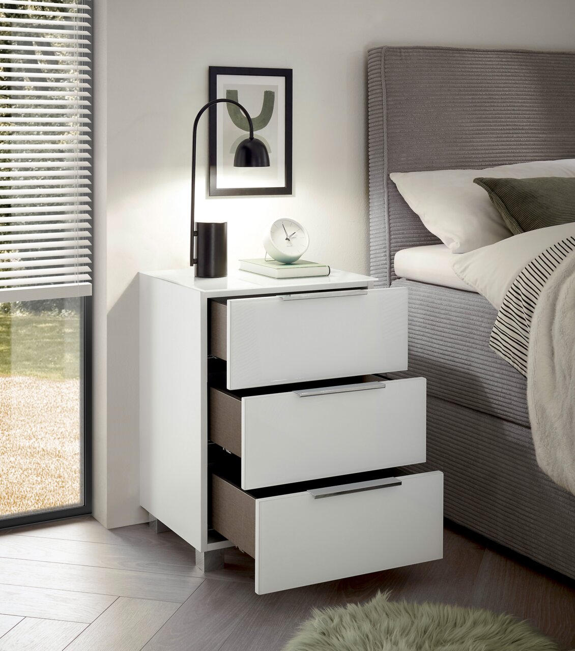 Moderne weiße Nachtkommode mit drei offenen Schubladen, seitliche Perspektive, dekoriert mit einer Lampe, einem Buch und einer Uhr, neben einem Bett.