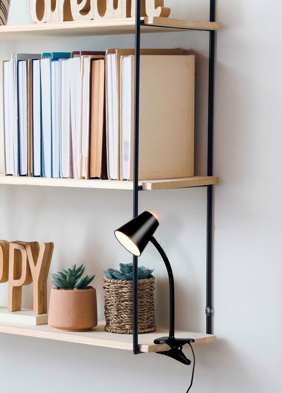Reality Leuchten Klemmleuchte PONGO Schwarze Klemmleuchte an einem Regalbrett, umgeben von Büchern und Pflanzen, seitliche Perspektive.