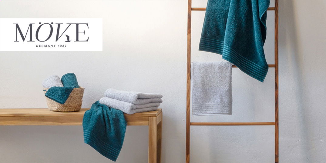 Weiche Handtücher in Türkis und Grau liegen gefaltet auf einer Bank und hängen an einer Holzleiter.