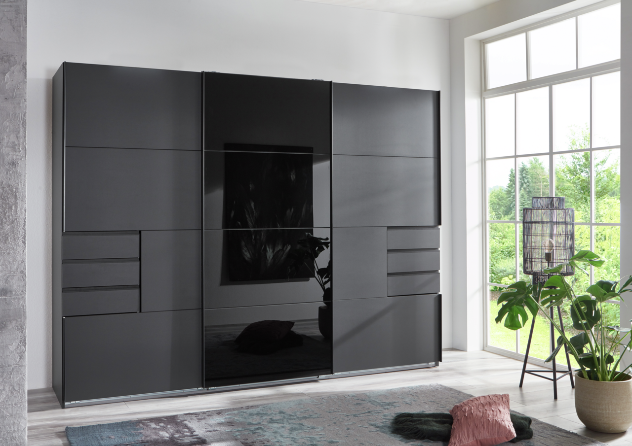 Z2 Schwebetürenschrank 3trg. Saigon Moderner, dreitüriger Schwebetürenschrank in dunklem Design, frontal abgebildet in einem hellen Raum mit großen Fenstern.