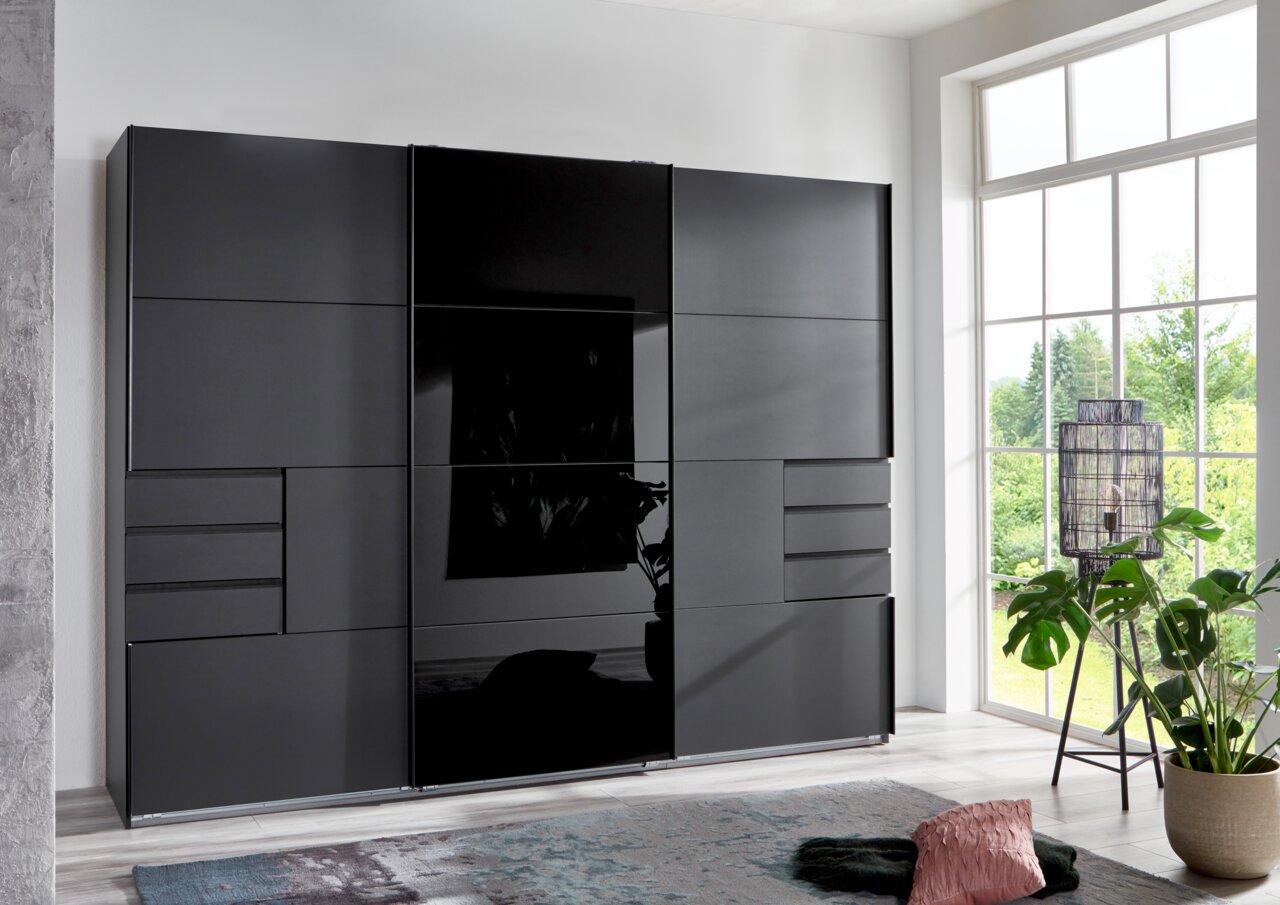 Moderner, dreitüriger Schwebetürenschrank in dunklem Design, frontal abgebildet in einem hellen Raum mit großen Fenstern.