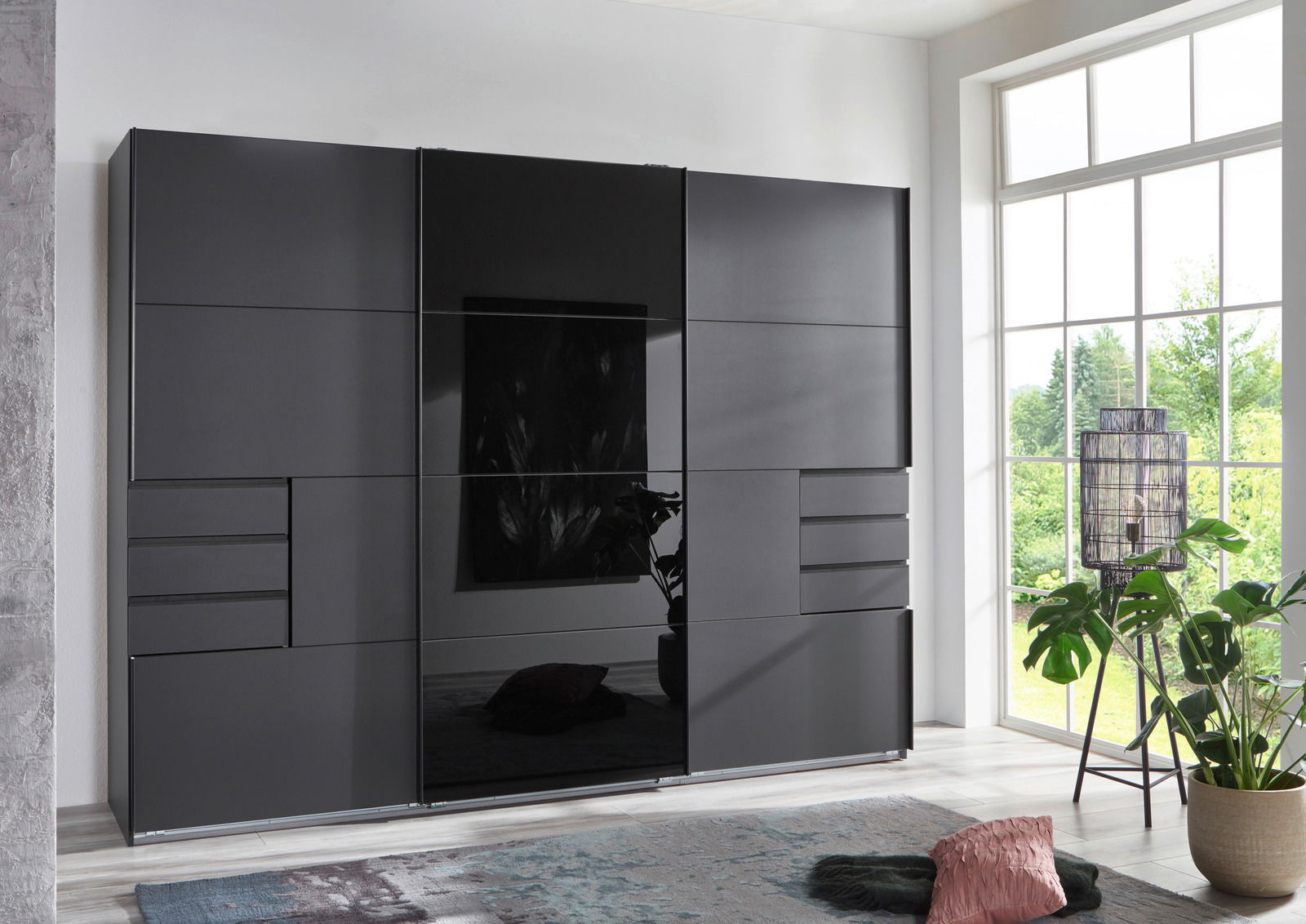 Moderner, dreitüriger Schwebetürenschrank in dunklem Design, frontal abgebildet in einem hellen Raum mit großen Fenstern.