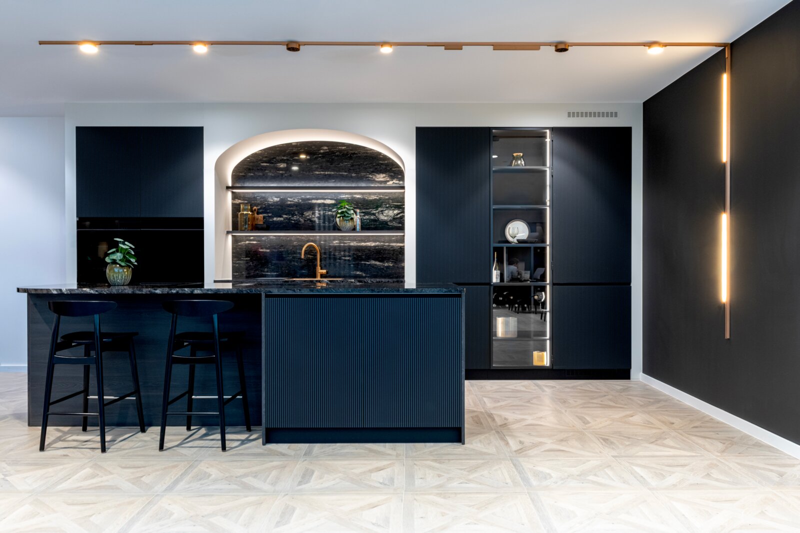 Frontalansicht einer modernen Designküche mit dunkelblauen, geriffelten Fronten, großer Kücheninsel mit zwei Barhockern, dunkler Naturstein-Arbeitsplatte und goldfarbener Armatur, beleuchtet durch Decken-Spotlights und vertikale Lichtleisten
